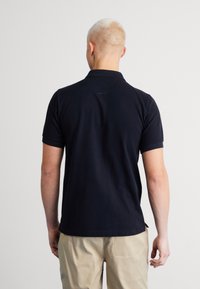 Polo de manga corta azul marino con cuello, textura suave y aberturas laterales, combinado con pantalones de color claro. Visto desde atrás.