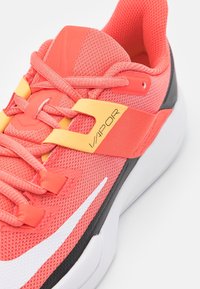 Korallfärgad mesh-idrottssko med gula accenter, vit swoosh och svart mellansula. Har plastdetaljer och platta snören för en säker passform.