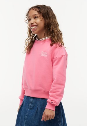 Uśmiechnięta dziewczyna o kręconych włosach nosi różową bluzę Tommy Hilfiger i niebieską spódnicę jeansową na jednolitym białym tle.
