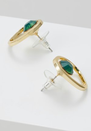 Boucles d'oreilles créoles en or avec pierres précieuses vertes facettées et supports en plastique transparent sur une surface blanche.