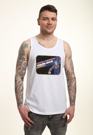 Biały bawełniany top na ramiączkach z grafiką postaci o imieniu Warlock w żywych kolorach na ciemnym tle. Szare dżinsy i widoczne tatuaże.