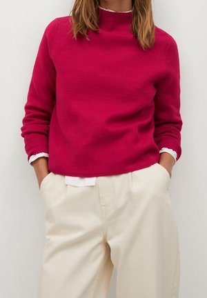 Kvinde iført en lyserød langærmet sweater over en hvid skjorte, der er stoppet ned i højtaljede cremefarvede bukser med hænderne i lommerne.