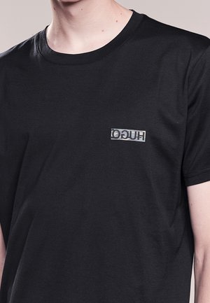 Person wearing a black short-sleeve T-shirt with a small, reversed "HUGO" logo printed on the left chest area.