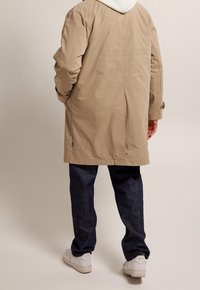 Beżowy długi płaszcz o gładkiej fakturze, z białym kapturem i bocznymi kieszeniami, zestawiony z ciemnoniebieskimi dżinsami i białymi trampkami.