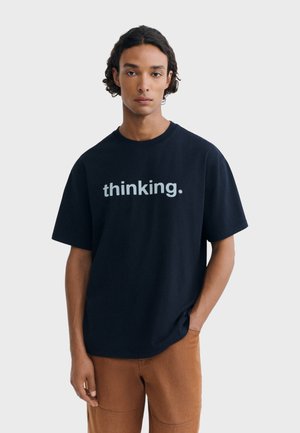 Homme aux cheveux bouclés portant un t-shirt noir avec le texte "thinking." et un pantalon marron, debout avec une main dans la poche devant un fond uni.