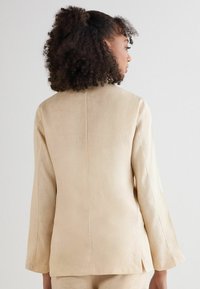 Beige linnen blazer met een getailleerde pasvorm, ronde halslijn en lange mouwen. Heeft een middelste rugnaad en subtiele zijsplitten.
