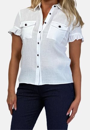 Femme portant un chemisier blanc à manches courtes avec des poignets à volants et un pantalon noir à pois, debout les mains détendues.