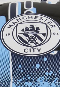 Cirkulært Manchester City-logo med et sort skib på et stribet skjold, omgivet af tekst. Baggrunden er blå med splatter og halftone-mønstre.