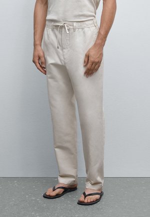Light beige linen pants with an elastic waistband and drawstring. Straight leg cut, smooth texture, worn with black flip-flops.