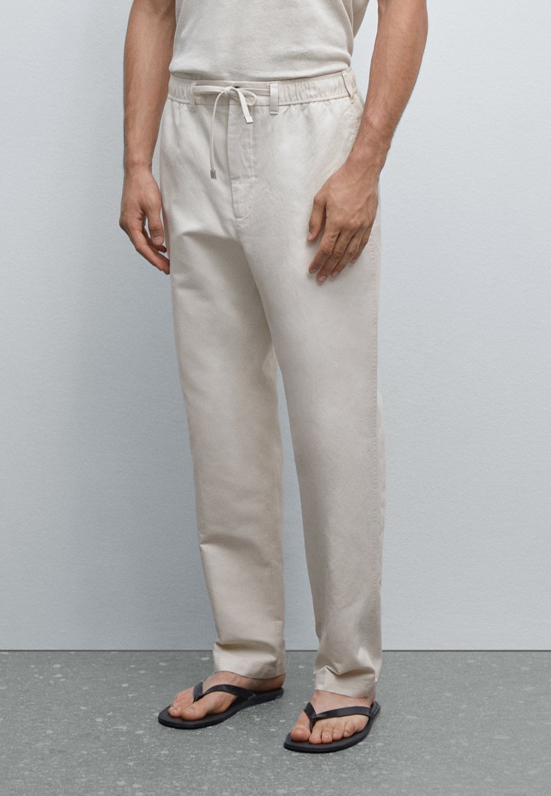 Ljusbeige linbyxor med resårlinning och dragsko. Rakt ben, slät textur, använda med svarta flip-flops.