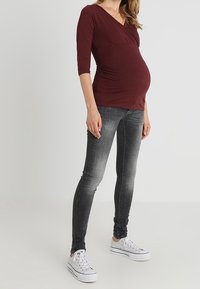 Těhotná žena v burgundském topu, vybledlých černých skinny džínách a bílých teniskách stojí proti jednoduchému světlému pozadí.