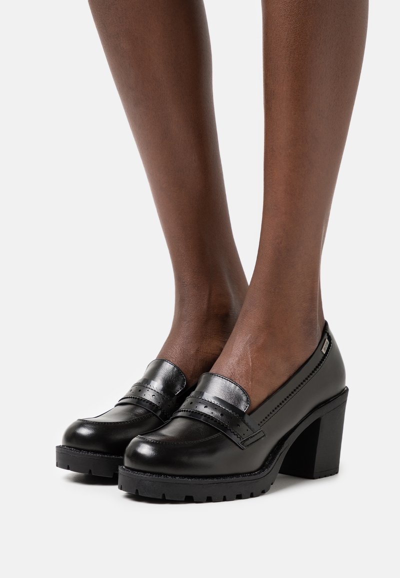 Escarpins en cuir noir avec une semelle épaisse, un bout rond et une bride décorative ornée de détails brogues. Texture lisse et design robuste.