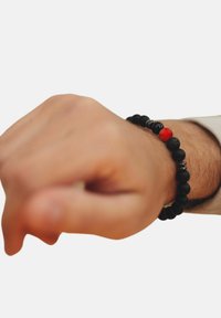 Pulsera de cuentas negras con una cuenta de acento roja, con una textura suave y formas redondas, llevada sobre un puño cerrado contra un fondo blanco.