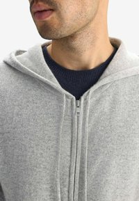 Ljusgrå zip-up hoodie i mjukt tyg, med dragsko i huvan och synliga sömnadsdetaljer, lagd över en marinblå tröja.