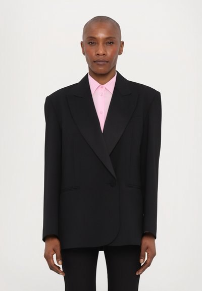 Schwarzer übergroßer Blazer mit Revers und einem einzigen Knopfverschluss, kombiniert mit einem hellrosa Hemd mit Kragen. Glatter Stoff mit minimalen Details.