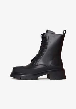 Schwarze Leder-Stiefeletten mit Schnürdesign, dickem Plateau und gerillter Laufsohle. Verfügt über eine Zuglasche an der Rückseite für ein einfaches An- und Ausziehen.