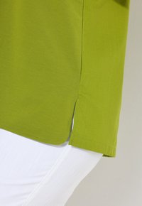 Camicetta verde a maniche corte con texture liscia e spacchi laterali, abbinata a pantaloni bianchi che mostrano sottili linee di tessuto.
