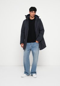 Marineblå polstret jakke med hætte, lynlåslukning og sidelommer, båret over en sort sweater og lyseblå lige jeans. Hvide sneakers.