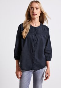 Camicia blu navy con bottoni e maniche a tre quarti, caratterizzata da pieghe orizzontali sul petto e un orlo arrotondato. Tessuto dalla texture liscia.