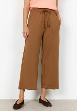 Žena stojaca pri bielej stene, oblečená v hnedých širokých krátkych nohaviciach s nastaviteľným pásom, broskyňovom sleeveless top a tmavohnedých plochých topánkach.