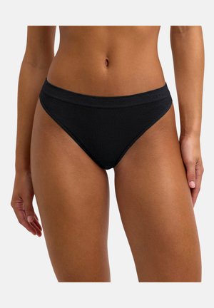 Femme portant un bas de bikini noir uni, vue de la taille jusqu’aux cuisses sur un fond uni clair.