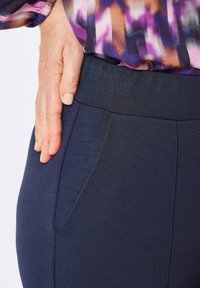 Donkerblauwe broek met een gladde structuur, voorzien van een hoge tailleband en een getailleerd ontwerp. Zijzak zichtbaar nabij de tailleband.