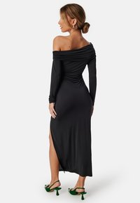 Svart off-shoulder klänning med långa ärmar och sidoklyfta, med draperad design och slät tygtextur. Kombinerad med gröna klackskor.