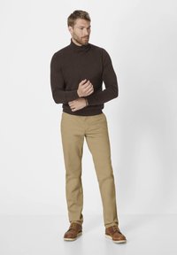 Brauner Rollkragenpullover, beigefarbene Hose, braune Lederschuhe. Mann steht mit verschränkten Armen, lässiger Schnitt, glatte Stofftextur.