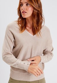 Pull en tricot beige avec un décolleté en V. Présente des accents de boutons sur les manches et un tissu doux et texturé. Coupe décontractée.