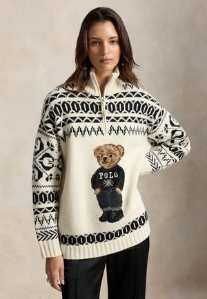 Mujer que lleva un suéter de punto blanco con patrones negros y un gráfico de un osito de peluche, combinado con pantalones anchos negros y zapatos negros.