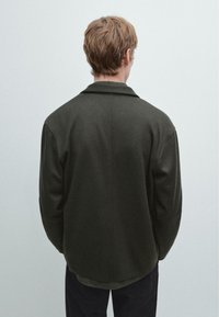 Persona con capelli corti castano chiaro che indossa una giacca verde scuro a maniche lunghe e pantaloni neri, vista di spalle su uno sfondo chiaro e uniforme.