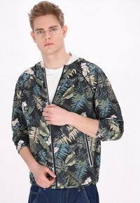 Giacca con cappuccio caratterizzata da una base nera con stampa floreale tropicale e foglie multicolore, chiusura con zip e maniche raglan.