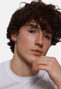 Modèle avec des cheveux bruns bouclés, portant une chemise blanche simple, a une boucle d'oreille noire dans l'oreille gauche ; la peau semble lisse et naturelle.