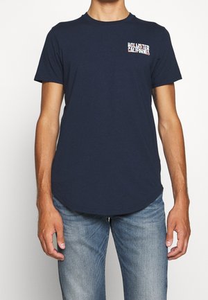 Marineblaues Rundhals-T-Shirt aus Baumwolle, mit kleinem grafischem Logo auf der linken Brust. Kurze Ärmel und eine geschwungene Saumlinie.