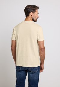Beige tričko s krátkým rukávem a kulatým výstřihem s hladkým povrchem, nošené s džíny, viděné zezadu.