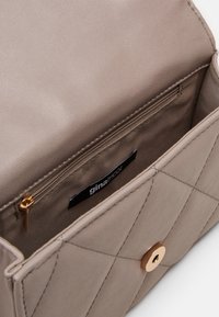 Borsa a tracolla trapuntata color taupe con chiusura dorata, aperta per mostrare la tasca con zip interna e l'etichetta in tessuto con la scritta "gina tricot."