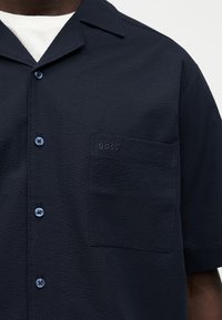 Κοντομάνικο μπλε πουκάμισο με υφή υφάσματος, τσέπη στο στήθος με κέντημα "BOSS", φορεμένο πάνω από λευκό φανελάκι.