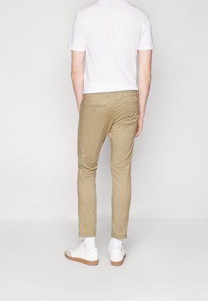 Chinos khaki avec deux poches arrière et quincaillerie en argent, associés à un t-shirt blanc uni et des baskets blanches. Design ajusté.