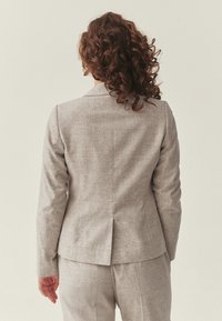 Mujer con pelo rizado y castaño, vestida con un traje a medida gris claro, de pie de espaldas contra un fondo liso y claro.