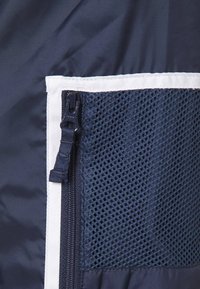 Gros plan d'une poche en maille bleu marine avec une fermeture éclair noire sur un tissu sombre, bordée d'un liseré blanc.