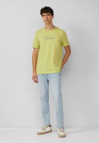 Żółta koszulka z krótkim rękawem i okrągłym dekoltem z napisem "S.Oliver Elevated Casual". Połączona z jasnoniebieskimi dżinsami i białymi trampkami z beżowymi akcentami.