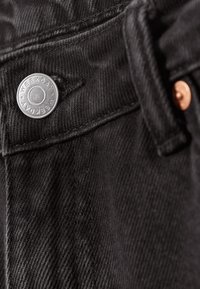 Tissu en denim noir avec une surface texturée, présentant un bouton circulaire en argent étiqueté "WEEKDAY" et un bouton-pression de couleur cuivre.