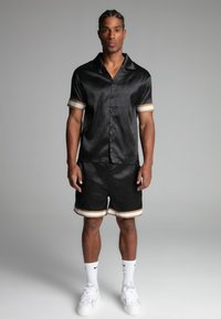Una maglietta nera a maniche corte con finitura lucida e polsini accentuati in oro, abbinata a pantaloni corti coordinati con rifiniture dorate. Indossata con sneakers bianche.