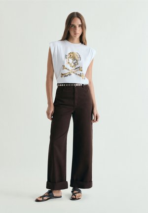 Tânără care poartă un top alb fără mâneci cu imprimeu aurit de craniu și oase încrucișate, pantaloni negri evazați, curea cu ținte și sandale negre.