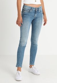 Jeans skinny de denim azul claro con un diseño desgastado. Presentan cinco bolsillos, pasadores para cinturón y un cierre de botón en la parte delantera. Combinados con zapatillas blancas.