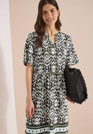 Femme en robe à motifs noir et blanc à manches courtes tenant un grand sac fourre-tout tissé noir, debout sur un fond beige.