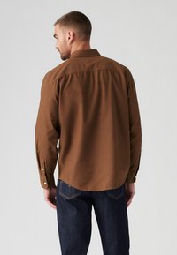 Brown long-sleeve shirt with button cuffs and a rounded hem, made of a smooth fabric. Worn with dark blue denim jeans.