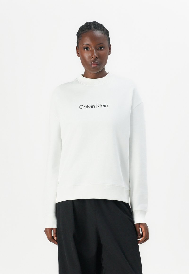 Biela mikina s rebrovaným výstrihom a manžetami, na prednej strane s čiernym logom "Calvin Klein". Kombinovaná s voľnými čiernymi nohavicami.