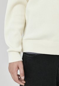 Pull en crème avec des côtes, doté d'une finition ajustée à l'ourlet et aux poignets, associé à un jean foncé. La texture est douce, avec un design simple et épuré.