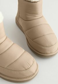 Stivaletti alla caviglia imbottiti beige con esterno liscio, suola testurizzata e fodera morbida. Presentano pannelli cuciti e design della punta arrotondata.